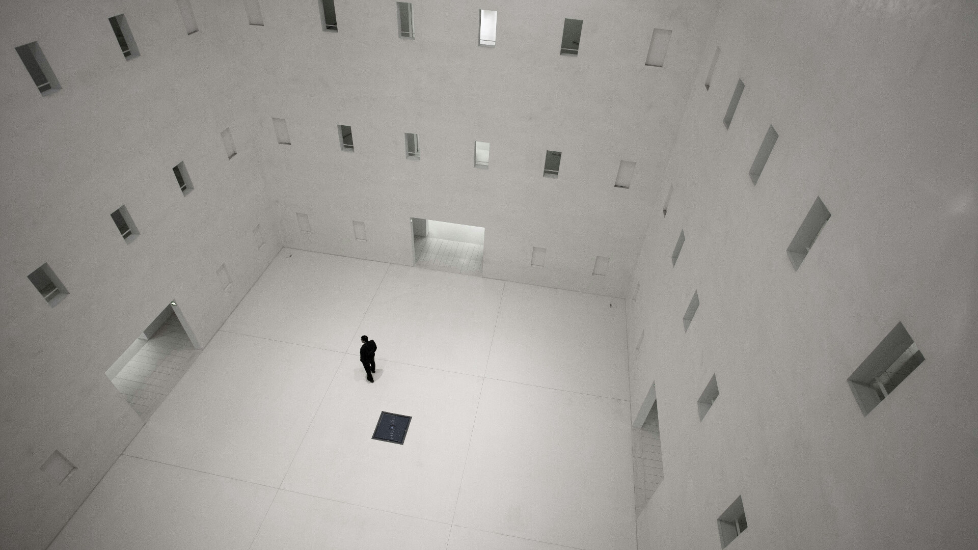 A lone figure standing in a vast, minimalist white atrium with grid-like windows